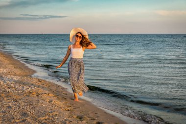 Beyaz şapkalı kadın gün batımında sahilde yürüyor.
