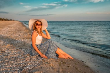 Beyaz şapkalı kadın gün batımında sahilde oturuyor.