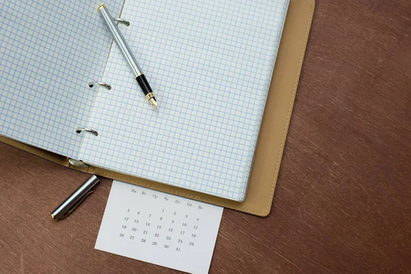 gümüş bir dolma kalem, mavi kapaklı bir defter ve ahşap bir masanın üzerinde bir takvim