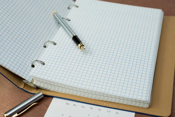 gümüş bir dolma kalem, mavi kapaklı bir defter ve ahşap bir masanın üzerinde bir takvim
