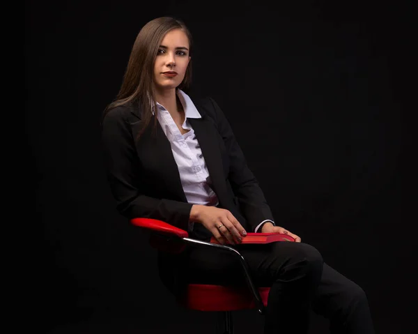 Genç ve güzel bir iş kadını. Sandalyesinde kırmızı bir not defteri ve koyu bir arka planı var. İş kıyafetleri içinde.