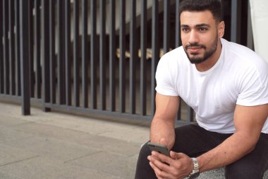 Şehir stadyumunun merdivenlerinde oturan genç adam. Akıllı telefon tutan temel beyaz tişört giymek ve ileriye bakmak. 