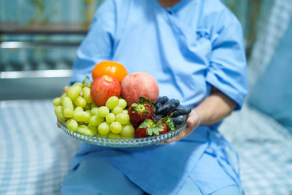 Asian senior or elderly old lady woman patient holding various fresh fruits healthy food with hope and happy while sitting and hungry on bed in hospital.