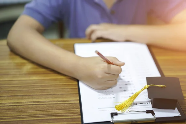 Asian children young kid write answer in a quiz examination test study learn with graduation gap hat on wooden table in school : bright genius education concept