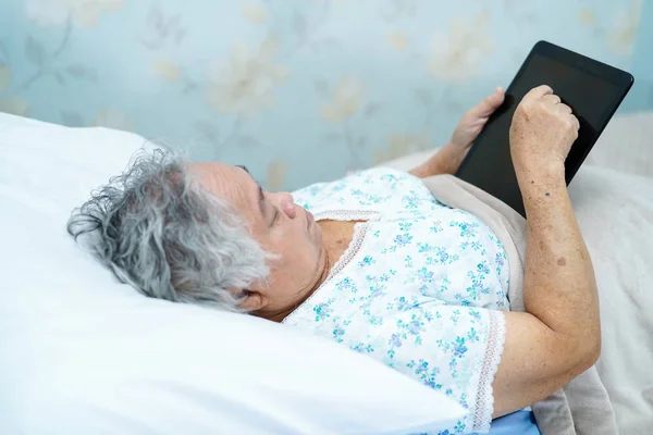 Asian senior or elderly old lady woman patient holding in her hands digital tablet and reading emails while sitting on bed in nursing hospital ward : healthy strong medical concept