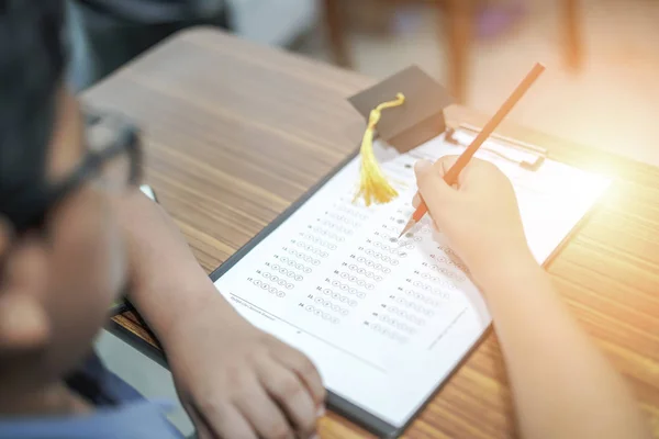 Asian children young kid write answer in a quiz examination test study learn with graduation gap hat on wooden table in school : bright genius education concept