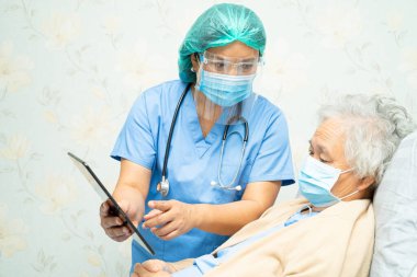 Doktor yüz kalkanı ve PPE giysisi giyiyor. Enfeksiyon hakkında veri toplamak, Roman Coronavirus 'u yaşlı bir kadınla öldürmek ve tedavi etmek için elinde dijital tablet tutuyor..