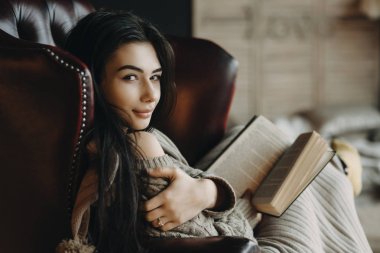 Esmer bir kitap okuma ve kameraya bakarak bir deri sandalyede oturan şaşırtıcı gülümseyen sol el ile sağ kolu dokunur.