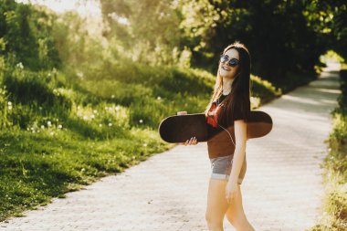 Onun elinde bir kaykay ile parkta yürüyüş ve kamera gülüyor içine bakarak güneş gözlüğü takıyor şaşırtıcı uzun saçlı kız.