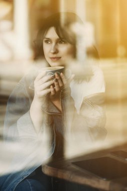 Portre çekici genç bir kadın pencereden bir fincan sıcak kahve içme ve uzak düşünceli arıyorum bir sandalye bir kafede oturmak.