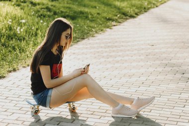 Tam uzunlukta bir kaykay üzerinde oturan ve smartphone açık arıyorum güzel bir beyaz kız portresi.