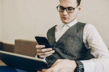 Kendine güvenen genç yönetici bir kanepe oturma ve bir tablet ile smartphone vasıl cidden bakarken işletim gözlüklü.