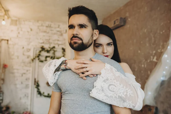 Cute caucasian couple where tattooed with red lips girl is embracing his men from back and looking into camera while her bearded man is looking away seriously.