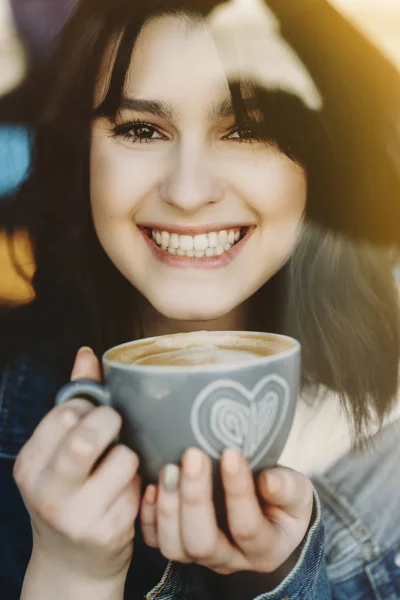 Close up portrait of a beautiful funny brunette drinking coffee and looking into camera laughing.