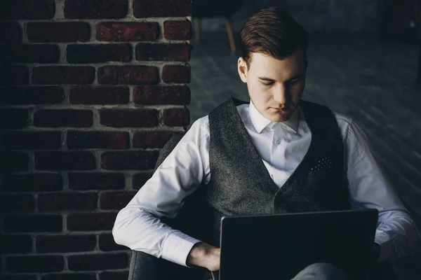 Confident elegant young executive operating on a laptop sitting in a chair near a window.