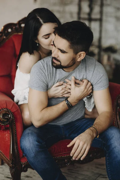 美丽的情侣 坐在红色的椅子上 微笑着在家里 而女孩是拥抱他的胡子丈夫从后面 — 图库照片