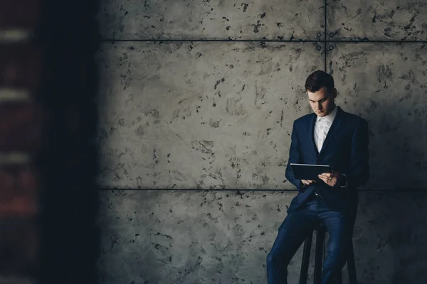 Handsome confident executive on a chair and operating with a tablet company\'s tasks against a grey wall.