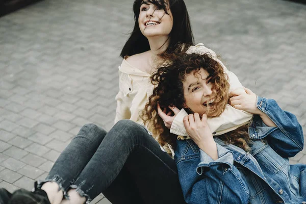 Charming two women having fun on the ground of a terrace in the city where brunette is holding her girlfriend\'s neck laughing while curly girl is looking into camera laughing.