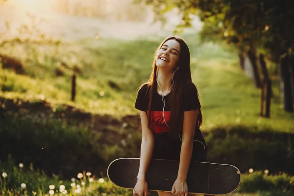 Beautiful caucasian girl having fun with a skateboard in her arms laughing and looking into camera in the park at the sunset.