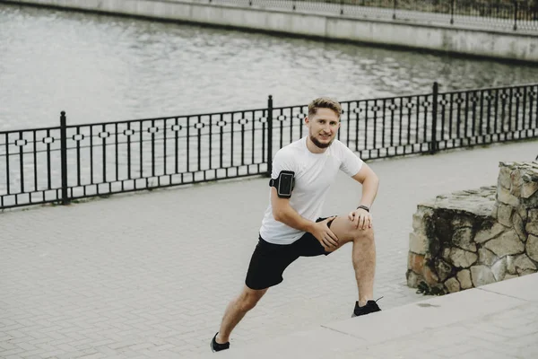 Young blonde man stretching outdoor in the morning looking into the camera and smiling against the park.