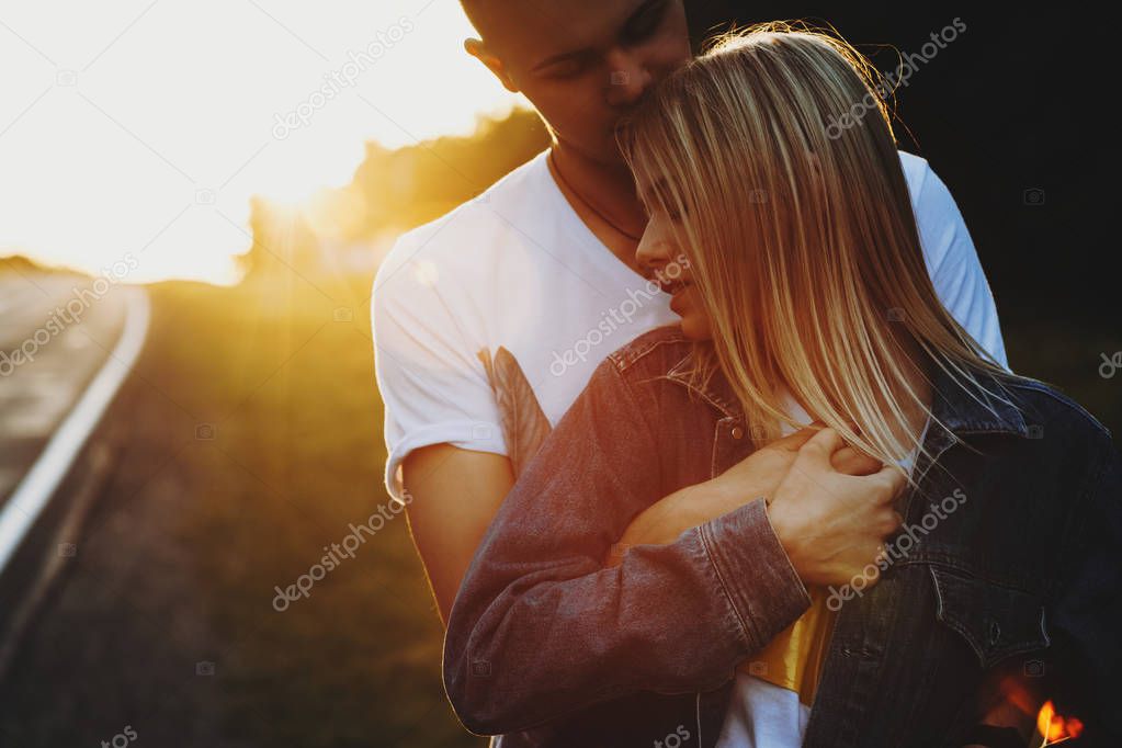 Close up portrait of a lovely caucasian couple reasting near the road in the forest where man is embracing his love from back while she is leaning her head on his chest with closed eyes smiling against sunrise.