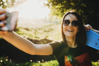 Güzel genç beyaz kız omuzlarını selfie karşı günbatımı gülüyor yapıyor güneş gözlüklü bir kaykay ile.
