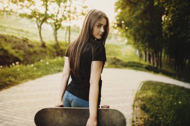 Büyüleyici bir uzun saçlı kız kamera karşı günbatımı gülümseyen bakarak ve bir kaykay tutarak geri gelen portre.