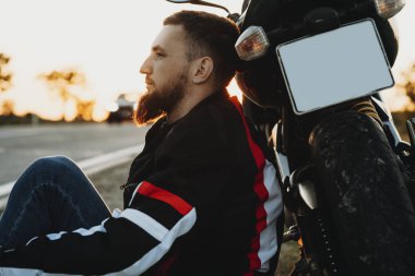 Bisikletiyle cidden yaslanmış ve uzak yola yakın motosikletine serüven dolu iken dinlenme seyir yerde oturan bir genç sakallı adam yan görünüm.