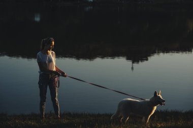 Onun beyaz Sibirya husky ile göl kenarındaki parkta günbatımında bir uzun saçlı beyaz kız yan görünüm.