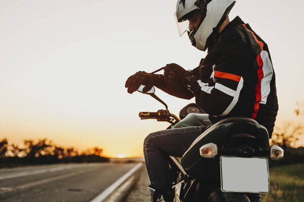 Side view of a caucasian man sitting on the motorcycle and putting his gloves preparing to start his adventure on motorcycle against sunset.