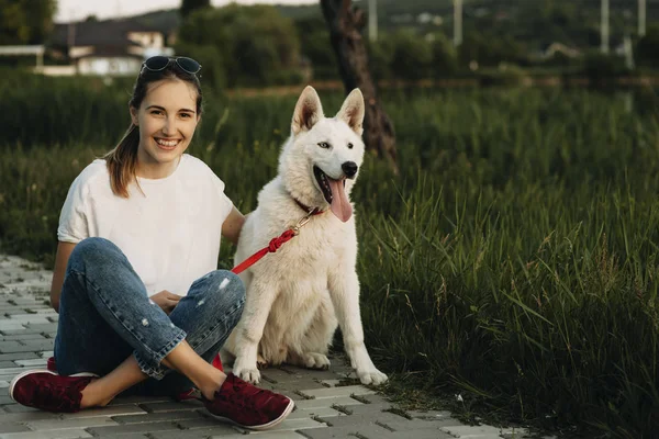 Happy cheerful female with crossed legs sitting on pavement hugging big white dog and looking at camera