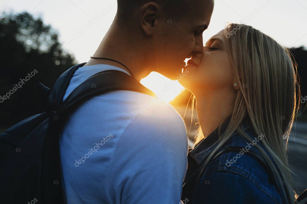Crop side view of smiling man and woman tenderly kissing standing at empty country road on backlit background with sun setting between them