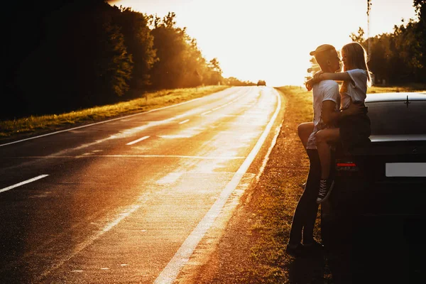 Side view of beautiful woman sitting on car trunk passionately embracing man holding hands around her waist on backlit background of empty road and trees in evening