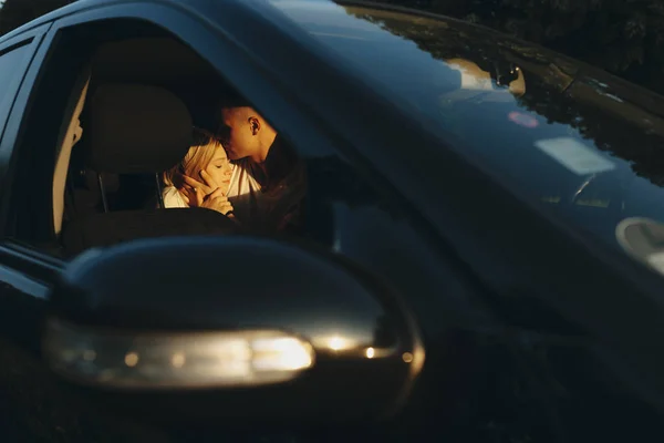 Adorable young couple sitting on car backseat, male kissing forehead of female with closed eyes shot made through open front car door window at sunset