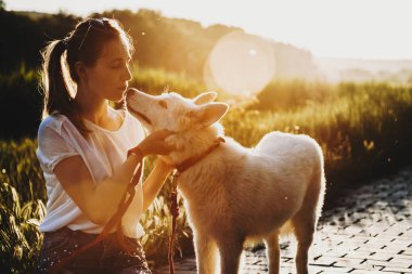 Yaz giysileri köpeğini arkadan aydınlatmalı arka plan üzerinde gün batımında yakın duran, kaldırım ve öpüşme burun kapalı gözler ile oturan at kuyruğu ile çekici genç bayan 
