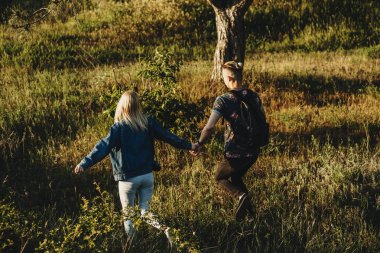 Arkadan görünüşü elele ve birlikte çalışan yeşil çayırlarda günbatımında yaz rahat kıyafetler içinde genç çift