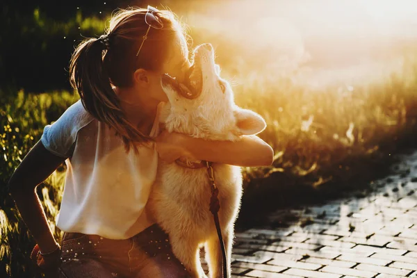 Akşam rahat yaz giysileri park kaldırım kadındaki kaldırdı baş ve bulanık arkadan aydınlatmalı arka plan üzerine açık çene ile sevimli beyaz köpek kucaklayan güneş gözlüğü ile oturuyor