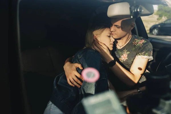 Handsome young man in summer clothes embracing delicately beautiful relaxed blond woman covered with jeans jacket leaning on him sitting on front seat of car with closed eyes