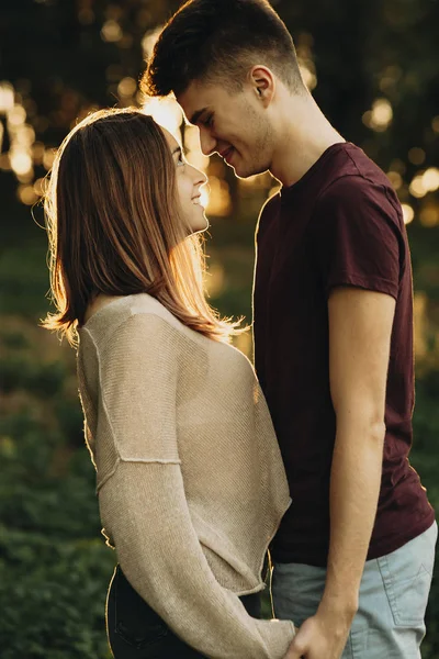 Side view of casual young man and woman holding hands and looking at each other standing in back lit in park