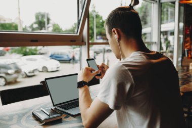 Yan görünüm genç adam oturmuş ve laptop kafede sayaçta smartphone kullanarak 
