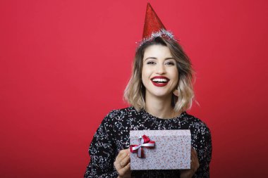 Parlak makyaj Noel hediyesi kapak kırmızı zemin üzerine kameraya bakıyor ve yayla sarılı holding ile şenlikli şapkalı gömlek ve kırmızı genç kadın