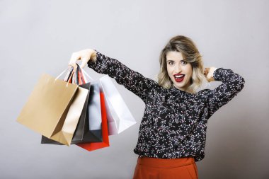 Sarı saçlar ve kağıt poşetler ile poz ve gri arka plan üzerinde kameraya bakarak bluz ve pişmiş toprak pantolon kırmızı ruj ile genç gülümseyen kadın