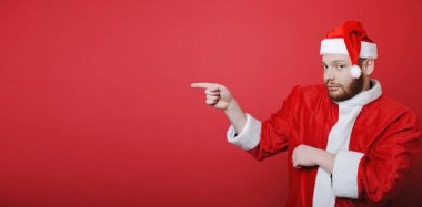 Kameraya bakarak ve bir kenara duran parlak kırmızı zemin üzerine işaret Noel Baba kostüm sakallı yakışıklı