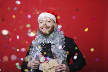Yakışıklı olan Noel şapka ve hediye tutarak ve başparmak-up jest düşen konfeti arkasında dururken gösterilen parlak tinsel parlak kırmızı zemin üzerine