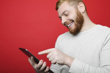 Gülümseyen ve parlak kırmızı bir arka plan üzerinde dururken modern tablet tarama kızıl sakallı yakışıklı genç adam