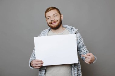 Gülümseyen ve kırmızı arka plan üzerinde duran ve boş poster işaret ederken kameraya bakarak Kızıl saçlı yakışıklı