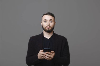 Yakışıklı olan smartphone kullanarak ve gri arka plan üzerinde dururken sürpriz yüz ifadesi ile kameraya bakarak şık kıyafeti