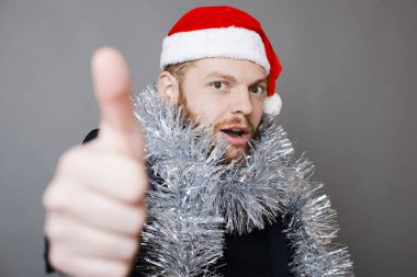 Genç adam Noel şapka ve kameraya bakarak ve başparmak-up jest duran gri arka plan üzerinde gösterilen parlak tinsel zencefil