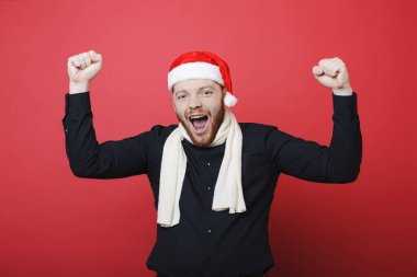 Fularlı çığlık ve parlak kırmızı arka plan üzerinde duran ve zaferini kutluyor kameraya bakarak Noel şapka ve sıcak yakışıklı sakallı adam