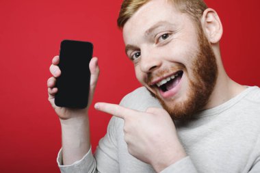 Smartphone boş ekran ile işaret ve parlak kırmızı bir arka plan üzerinde dururken heyecanlı yüz ifadesi ile kameraya bakarak kızıl sakallı yakışıklı adam